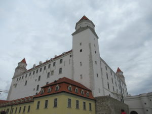 The Bratislava Castle is a large, white castle. Three towers are visible in the photo. In the bottom left corner of the photo, beneath the castle, is a yellow building with a red roof.