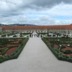 Image is of a baroque garden, with a wide lane down the middle of the picture. On either side are short bushes and flowers planted to make a design. On the far side of the picture there is a long building, with a red roof and yellow walls.