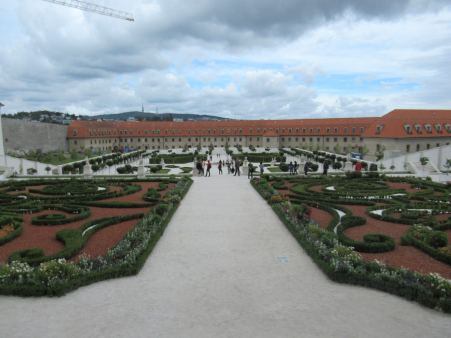 Image is of a baroque garden, with a wide lane down the middle of the picture. On either side are short bushes and flowers planted to make a design. On the far side of the picture there is a long building, with a red roof and yellow walls.