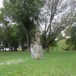 There is a grassy park area, with various trees in the background. In the center is a bronze statue of a woman with a man at her feet on a stone pedestal. Beside the pedestal is a bronze scroll describing who she is.