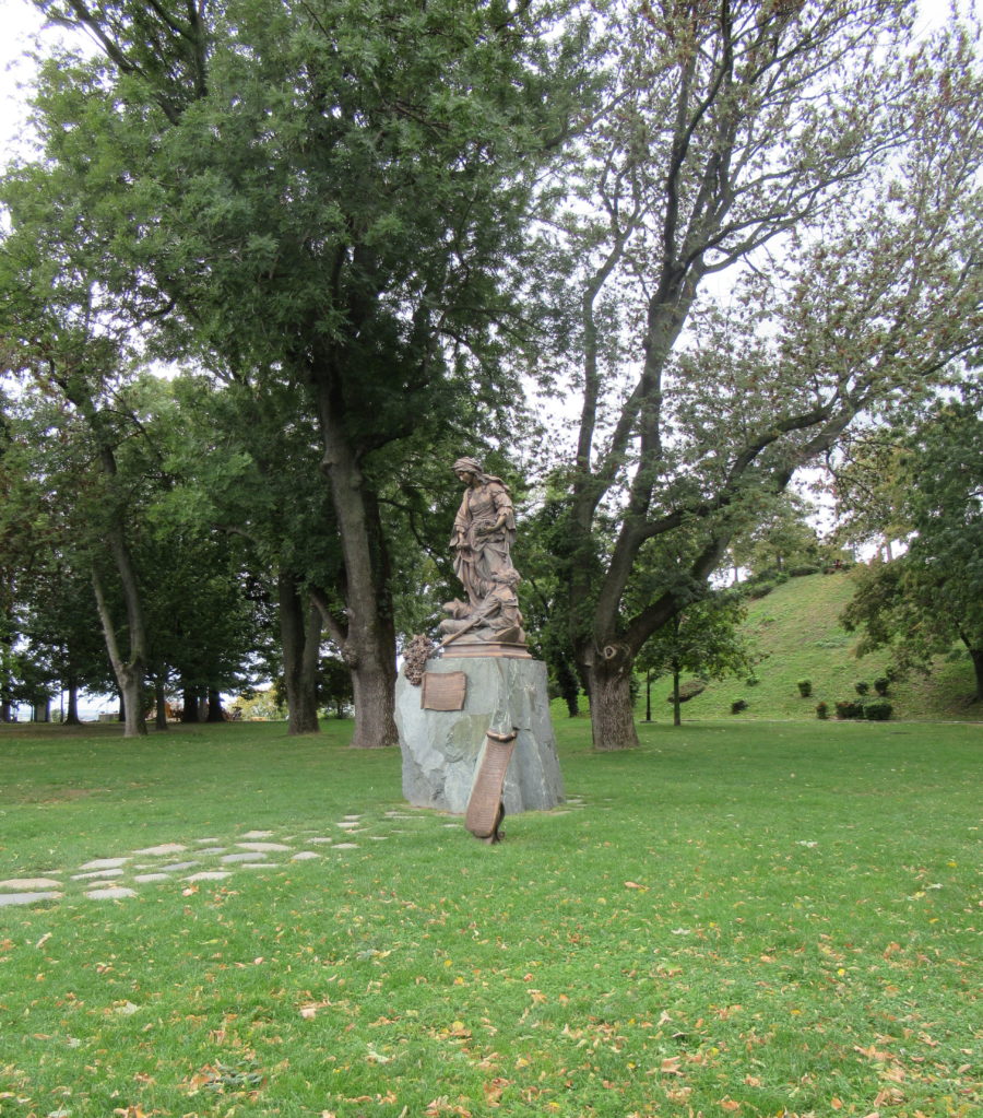 There is a grassy park area, with various trees in the background. In the center is a bronze statue of a woman with a man at her feet on a stone pedestal. Beside the pedestal is a bronze scroll describing who she is.