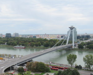 There is a bridge going over the Donau River. On the far side of the bridge there are two slanted pillars that support a circular, UFO-shaped building.