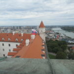 The Bratislava Castle viewed from a tower. Two other towers are visible, and from the roof of the castle, the Slovakian flag is unfurled. Behind the castle and to the right there is the city of Bratislava, and the Donau River.