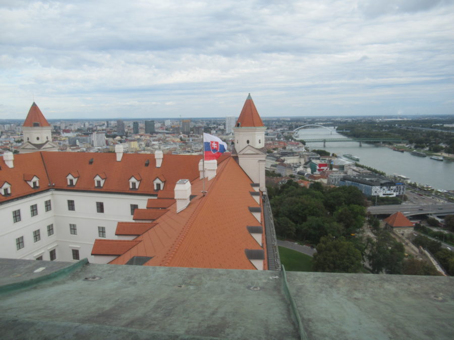 The Bratislava Castle viewed from a tower. Two other towers are visible, and from the roof of the castle, the Slovakian flag is unfurled. Behind the castle and to the right there is the city of Bratislava, and the Donau River.