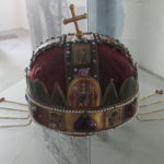 A domed crown, with a golden metal around the bottom, red velvet panels, and jewels. At the top is a golden cross, slightly askew. Hanging off each side of the crown are golden chains with red stones.