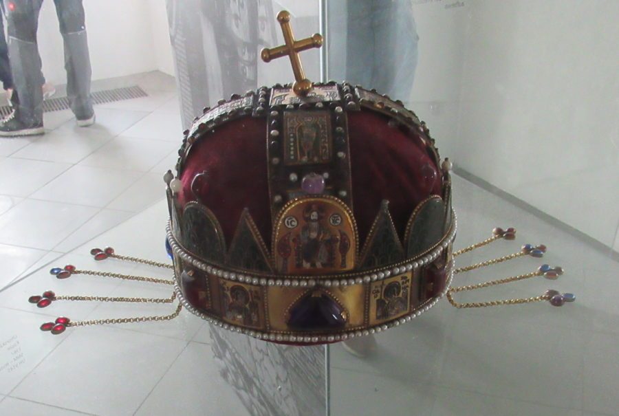 A domed crown, with a golden metal around the bottom, red velvet panels, and jewels. At the top is a golden cross, slightly askew. Hanging off each side of the crown are golden chains with red stones.