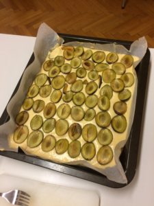 A pan with an uncooked cake. There are plums, halved and pitted, laid in rows on top of the batter.