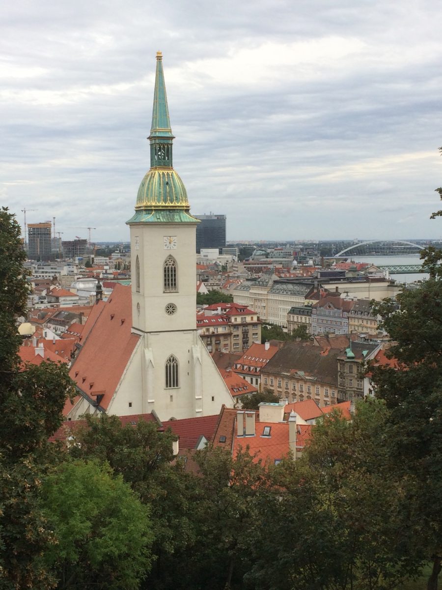 Image is of a church framed by trees along the bottom. The building is white, with a red roof, and the spire is green with gold accents. Behind the church are similar red-roofed buildings.